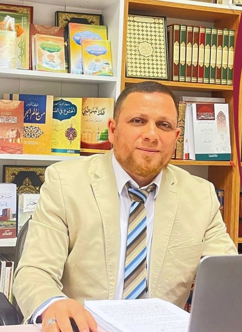 Man in beige blazer and striped tie sitting at desk with laptop and papers, bookshelves with Arabic books visible behind him
