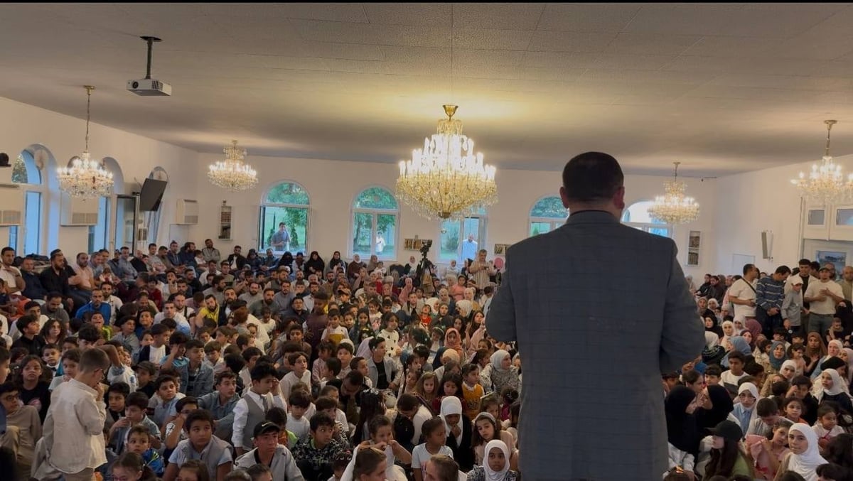 Man in suit standing alone facing large crowd of people in elegant ballroom with crystal chandeliers and large windows
